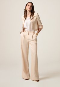 Blazer de piersici deschis, croit ajustat, cu mâneci trei sferturi, asortat cu pantaloni evazați cu talie înaltă. Bluză albă, netedă și strălucitoare; accesorii aurii.