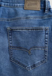 Los jeans de mezclilla azules cuentan con un solo bolsillo trasero con costuras en contraste y una etiqueta de cuero marrón que dice "Signature Denim Goods".