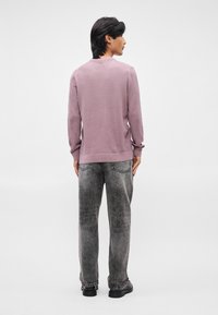 Lys lilla, struktureret sweater med lange ærmer; parret med slidte grå jeans og sorte sko. Bagfra set mod en ensfarvet baggrund.