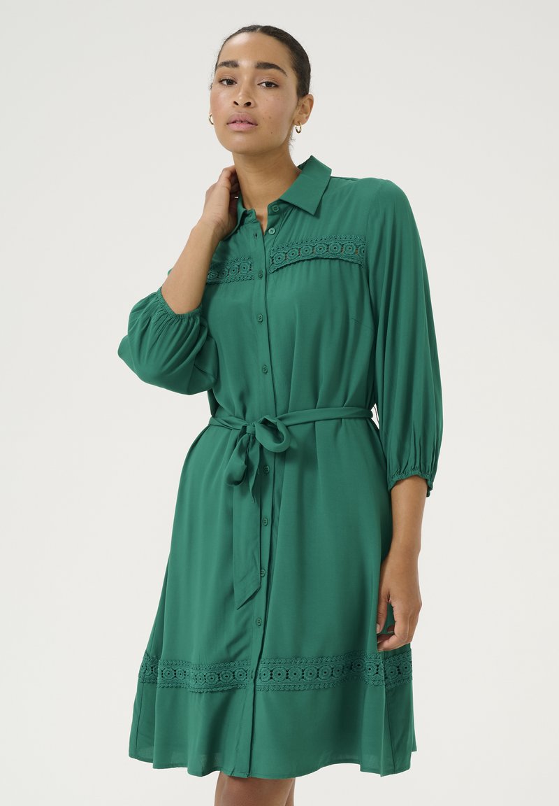 Robe verte à boutons avec une coupe décontractée, manches 3/4, ceinture à la taille et détails en dentelle le long du corps et de l'ourlet. Texture de tissu lisse.