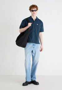 Camicia blu scuro con maniche corte e taschino, jeans dritti blu chiaro, borsa nera e occhiali da sole neri. Texture morbide.