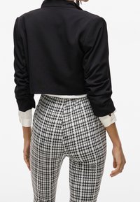 Femme portant un blazer noir court par-dessus une chemise blanche avec un pantalon taille haute à motif pied-de-poule, vue de dos.