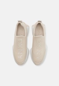 Beige slip-on sneakers med DKNY-merke, glatt syntetisk overdel, rund tå, hvit såle og teksturerte detaljer på hælen.