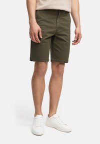 Pantalones cortos de algodón verde oliva con un frente plano, presillas para cinturón y dobladillo enrollado. Combinados con zapatillas blancas sobre un fondo liso.