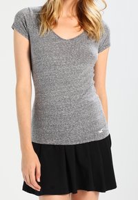 Grå V-ringad T-shirt i mjukt, melerat tyg, kombinerad med en svart plisserad kjol. Tröjan har korta ärmar och en diskret logotyp.