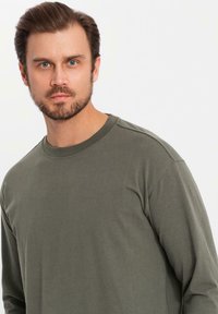 T-shirt a maniche lunghe in verde oliva, realizzato in tessuto morbido, con colletto a girocollo a coste e vestibilità rilassata. Dettagli delle cuciture sulle spalle.