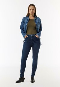 Jeansjacke in Hellblau, tailliertes olivgrünes Rippenoberteil, dunkelblaue Skinny-Jeans und schwarze Ankle-Boots mit spitzen Zehen.