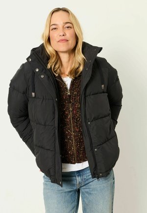 Blonde Frau trägt eine schwarze Steppjacke über einem braunen Strick-Zip-Pullover und hellblauen Jeans und steht mit den Händen in den Taschen.