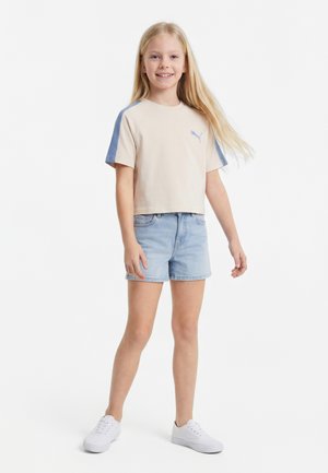 Jeune fille debout, portant un t-shirt court beige à rayures bleues, un short en jean clair et des baskets blanches, souriant sur fond blanc.