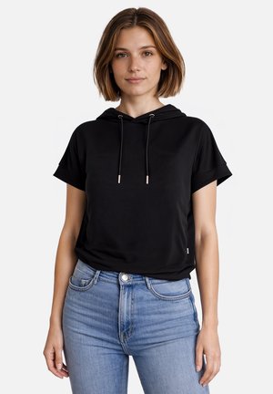 Jeune femme portant un sweat à capuche noir à manches courtes et un jean taille haute bleu clair, debout devant un fond blanc uni.