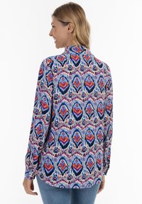 Blusa a maniche lunghe con motivo multicolore, collo a camicia e intricate decorazioni blu, viola e rosse su uno sfondo chiaro.