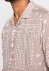 Camicetta a maniche corte, con bottoni, in tessuto marrone chiaro che presenta motivi paisley e geometrici bianchi, con una texture morbida e un design a colletto da campeggio.