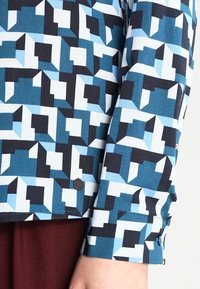 Long-sleeved shirt featuring a geometric pattern in shades of blue, black, and white. The fabric is smooth with a buttoned cuff detail.