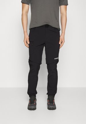Man die zwarte outdoorbroek met afritsbare korte broek draagt, donkergrijze wandelschoenen en een grijs T-shirt tegen een effen achtergrond.