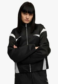 Giacca nera con zip, dettagli bianchi, collo alto e maniche larghe. Tessuto liscio, design corto, con pannelli laterali a contrasto.