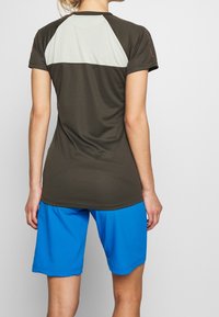 Krátký rukáv sportovního trička v olivově zelené barvě s světle šedou síťovinou na zádech, v kombinaci s modrými šortkami, hladká textura a uvolněný střih.