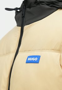 Μπεζ φουσκωτό μπουφάν με μαύρη κουκούλα, διαθέτει μπροστινό φερμουάρ και μπλε ετικέτα με το λογότυπο "HUGO". Ομαλή υφή, ζεστός σχεδιασμός.