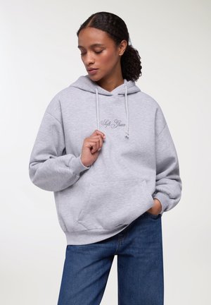 Felpa grigia chiara con coulisse regolabile, tasca frontale e ricamo "Soft Grace". Indossata con jeans blu, dal taglio casual.
