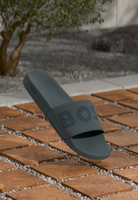 Dark gray rubber slide sandal with a wide strap featuring an embossed logo, set against a textured wooden tile surface.