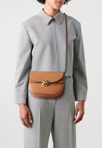 Brun lädercrossbodyväska med en rundad form, guldfärgade detaljer och ett klaffstängning. Används över en svart och vit rutig outfit.