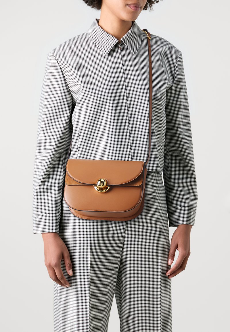 Brun lädercrossbodyväska med en rundad form, guldfärgade detaljer och ett klaffstängning. Används över en svart och vit rutig outfit.