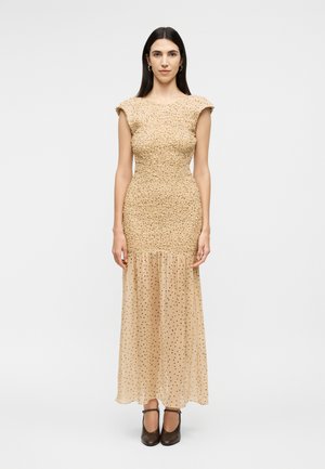 Γυναίκα με μακριά σκούρα μαλλιά, φοράει φόρεμα μπεζ με υφή, με διάφανο κάτω μέρος φούστας με πουά μοτίβο και μαύρες κλειστές γόβες, κοιτώντας προς τα εμπρός.