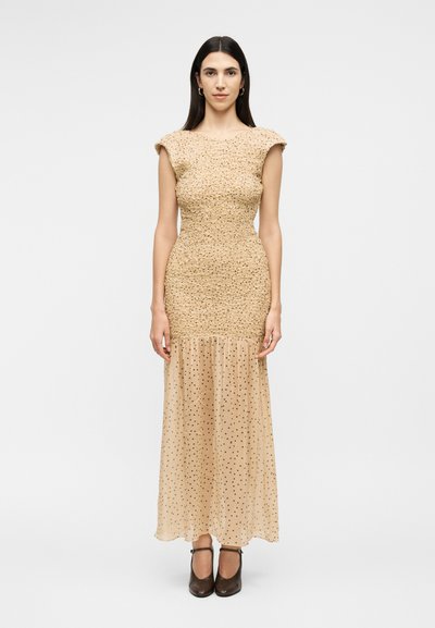 Femme aux longs cheveux foncés portant une robe beige texturée avec une jupe inférieure transparente à pois et des talons fermés noirs, faisant face vers l'avant.