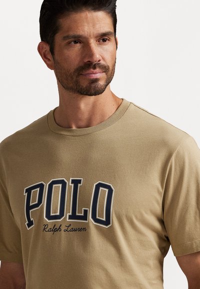Moški s kratkimi temnimi lasmi in brado, oblečen v bež majico z logotipom "POLO Ralph Lauren", ki gleda nekoliko na stran, na enobarvni belem ozadju.