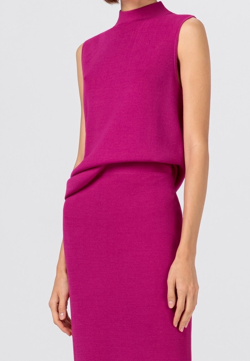 Tajt ärmlös klänning i klarröd färg, stickad tyg. Har hög hals och draperad detalj vid midjan, vilket skapar en smidig silhuett.