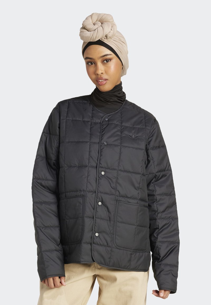 Femme portant un turban beige et une veste matelassée noire avec des boutons-pression et des poches avant, posant devant un fond clair.