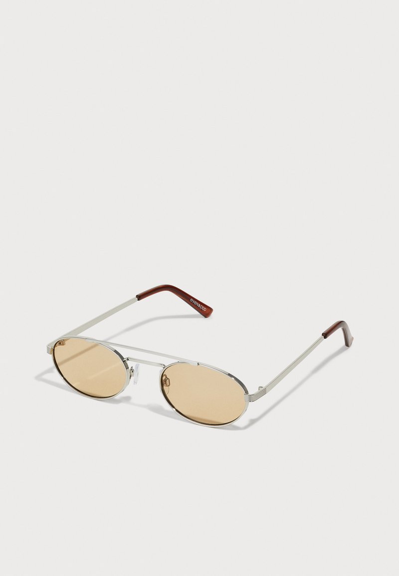 Lunettes de soleil en métal avec des verres ovales teintés de brun clair. Monture fine en argent avec des accents texturés et des embouts brunrouge sur les branches.