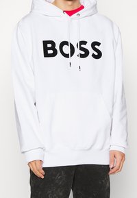 Weißer Kapuzenpullover aus weichem Stoff, mit großem schwarzem "BOSS"-Logo auf der Brust, einer Fronttasche und verstellbaren Kordeln.