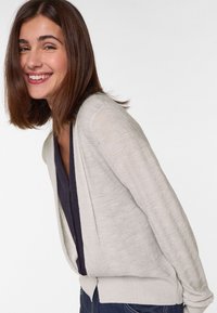 Lichtgrijze cardigan van textuurstof, open voorkant en lange mouwen, gedragen over een donkerblauwe top, gecombineerd met donkere jeans.