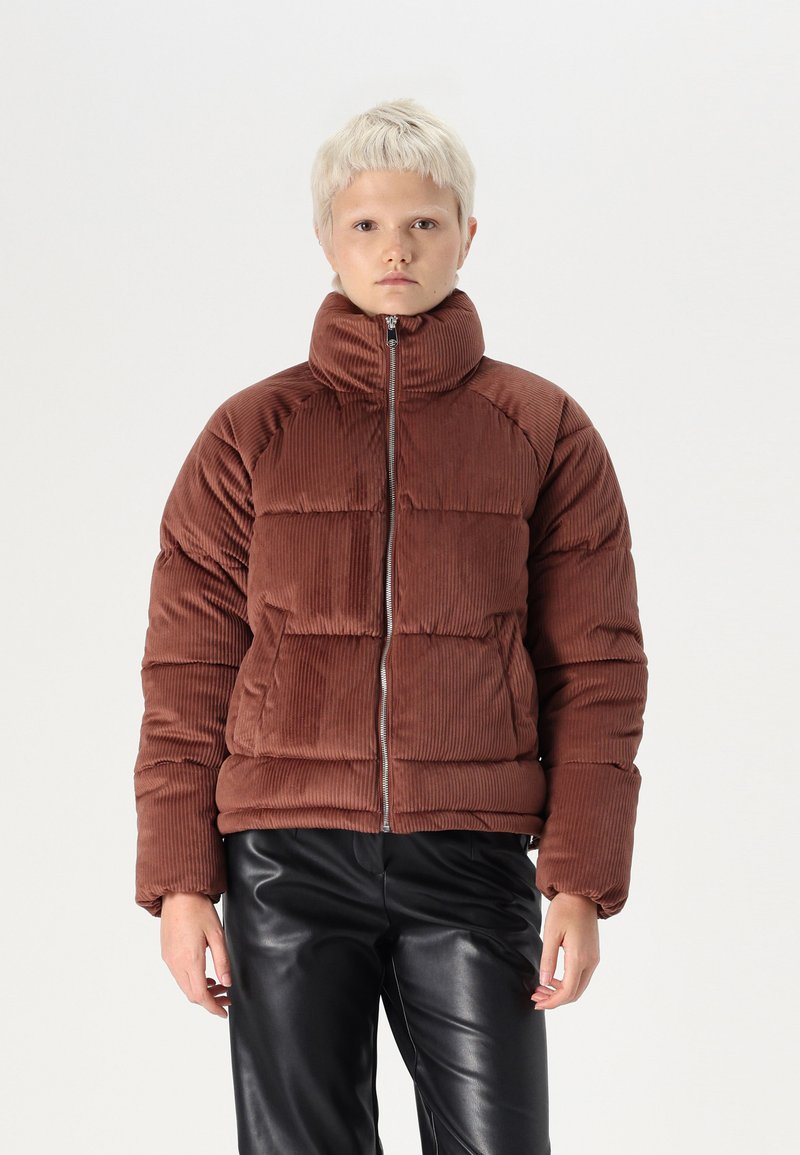 Brun fløyelsjakke med høy krage, glidelås foran og quiltet design. Kombinert med sorte skinnbukser.