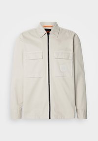 Jasno beżowa kurtka wykonana z bawełny, z zamkiem błyskawicznym z przodu, dwoma kieszeniami na piersiach oraz kwadratową naszywką z logo na lewej kieszeni.