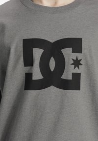 Pilkas medvilninis marškinėliai su dideliu juodu grafiniu dizainu, kuriame persipina raidės "DC" ir žvaigždutės akcentas. Minkšta tekstūra, apvali iškirptė.