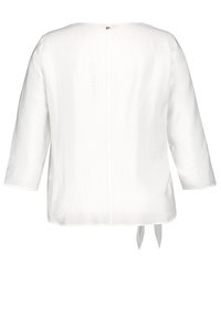 Blouse blanche à manches longues avec encolure ronde, texture froissée, détail de couture au dos, et deux liens en tissu suspendus à l'ourlet.