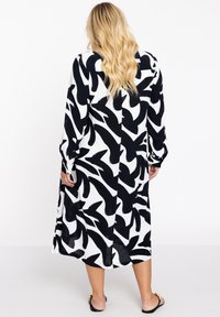 Schwarz-weißer Langarmkleid mit abstraktem Muster; weicher Stoff; lockere Passform; seitliche Schlitze; fällt unter das Knie; kombiniert mit schwarzen Sandalen.