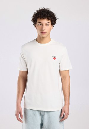 Jonge man met krullend haar die een effen wit T-shirt draagt met een kleine rode tomaatgrafiek en tekst op de linkerborst, gecombineerd met lichtblauwe spijkerbroek.