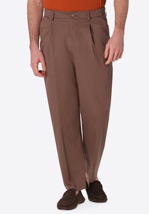 Uomo che indossa pantaloni marroni a pieghe e mocassini marroni scuri, abbinati a una camicia arancione, in piedi con i piedi leggermente divaricati.