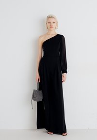 Svart en-skulder jumpsuit med lang gjennomsiktig erme, tett bodice, vid benbukse og en veske med geometrisk mønster. Jevn stoff tekstur.