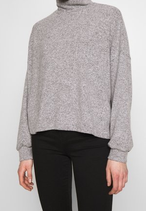 Pull à col roulé gris clair à manches longues, coupe ample, porté avec un pantalon noir sur une personne debout.