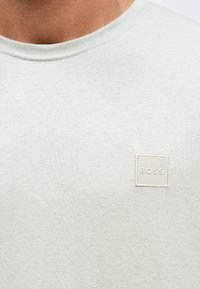 Ανοιχτό γκρι μπλουζάκι από υφασμένο βαμβάκι, με στρογγυλή λαιμόκοψη και ένα μικρό, τετράγωνο μπεζ ταμπελάκι με το λογότυπο "BOSS" στο στήθος.