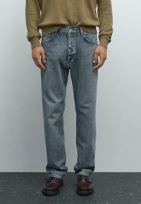 Uomo che indossa jeans azzurri, scarpe in pelle marrone e un maglione a maniche lunghe verde oliva, in piedi su un pavimento scuro a macchie.
