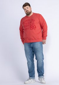 Rode sweatshirt met een print aan de voorkant, blauwe jeans en witte sneakers. De sweatshirt heeft een ronde hals en lange mouwen.