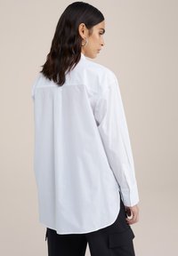 Camicia bianca a maniche lunghe in cotone, con piega sulla spalla posteriore, orlo arrotondato e polsi con bottoni. Tessuto morbido e vestibilità rilassata.