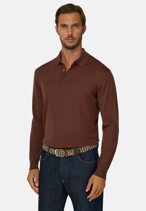 Hombre con camisa tipo polo marrón de manga larga, vaqueros azul oscuro y un cinturón tejido marrón claro y marrón, de pie frente a un fondo sencillo.
