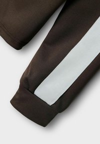 Mörkbrunt tyg med en strukturerad yta och vit horisontell randdetalj. Manschetten är fint sydd, vilket framhäver designkontrasten.