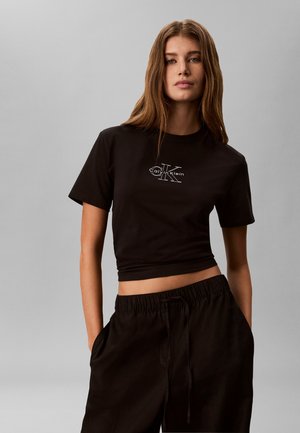 Mujer joven con cabello largo y castaño, que lleva una camiseta negra corta de Calvin Klein y pantalones negros, de pie frente a un fondo gris.