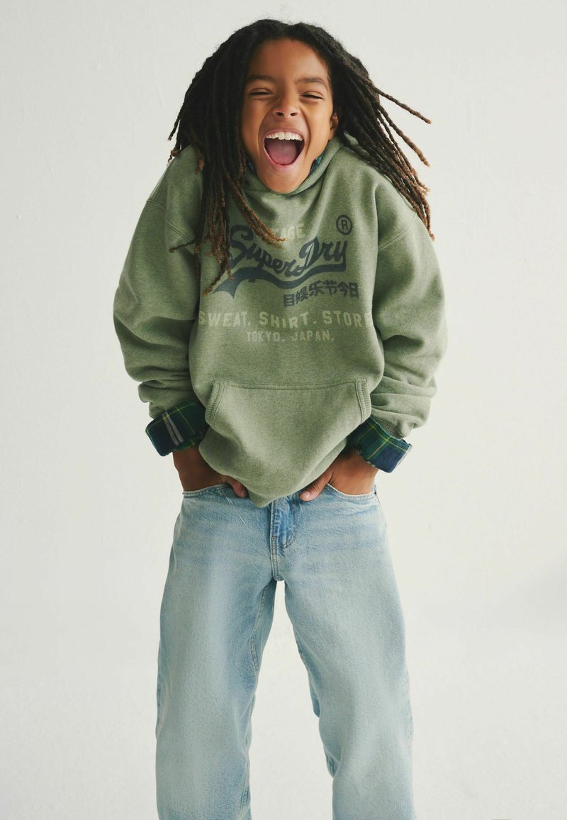 Sudadera verde con texto "Super Dry", jeans azules de corte holgado. La sudadera tiene una textura suave y las mangas presentan un detalle de cuadros en los puños.